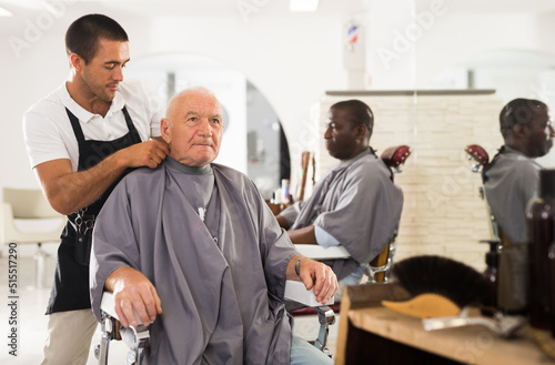 Professional stylist preparing aged man for haircutting, dressing hairdressing cape..