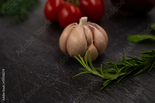 Close-up of a sprig of rosemary on a metallic dark background, with a head of garlic and other vegetables in the background.