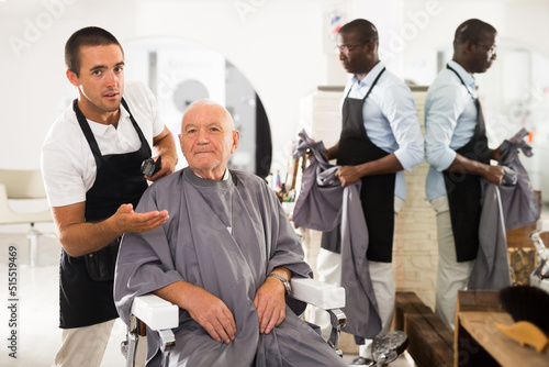 Smiling gray-haired man sitting in hairdressing chair, discussing haircut with young professional barber