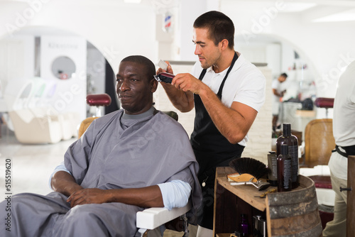 African-American man getting haircut with machine from skillful young barber in hair salon..