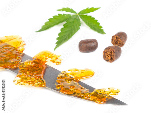 golden wax on a knife and a green cannabis leaf with pieces of hash on a white background.