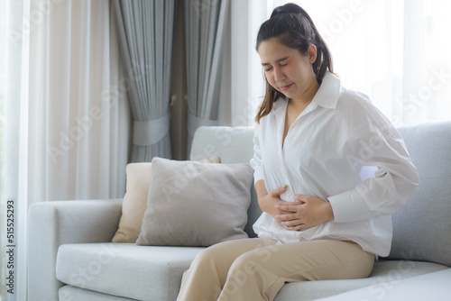 A woman wearing a white dress sat on the sofa in the living room with her hands clasped in her stomach. Her stomach ached so badly that she had to go to the hospital.