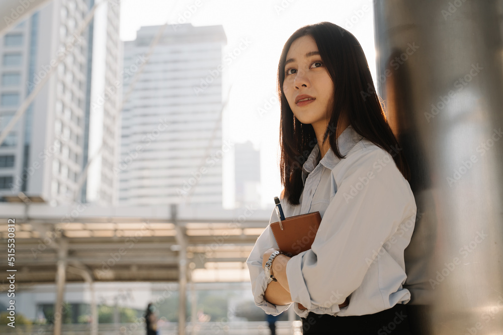 Portrait of beautiful young Asian woman standing leaning against building pillar holding a notebook and pen while thinking about something and looking away Portrait of beautiful young Asian woman standing leaning against building pillar holding a notebook and pen while thinking about something and looking away