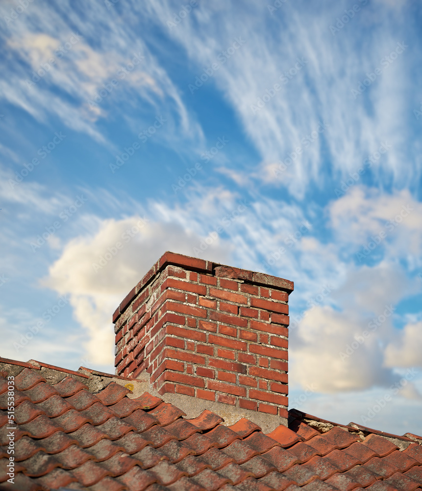 A brick chimney on the roof in the blue cloudy sky background. A closeup of the chimney on the rooftop. An orange bricked roof with chimney against the blue sky with copy space on a sunny day. A brick chimney on the roof in the blue cloudy sky background. A closeup of the chimney on the rooftop. An orange bricked roof with chimney against the blue sky with copy space on a sunny day.