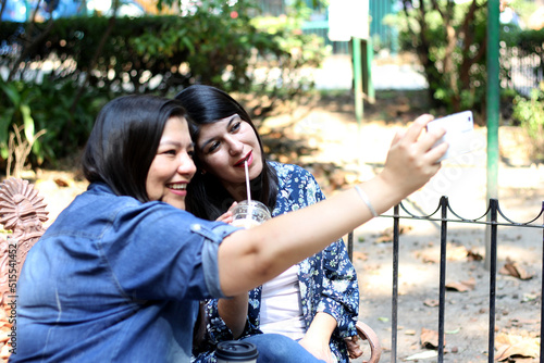 Two Latin women sister friends are sitting on a park bench outdoors using their cell phone to check messages, shop online and make love dates in app 