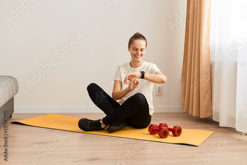 Full length athletic woman in white t shirt and tights sitting on yoga mat looking at her fitness tracker, checking indicators after workout, sitting with positive facial expression and smile.