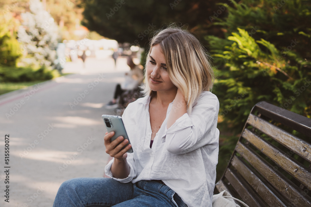 Charming young woman in casual clothes sits on a bench in the park and uses a mobile phone to communicate Charming young woman in casual clothes sits on a bench in the park and uses a mobile phone to communicate
