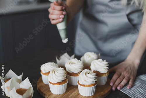 Young lovely woman pastry chef decorates cupcakes with whipped cream at home