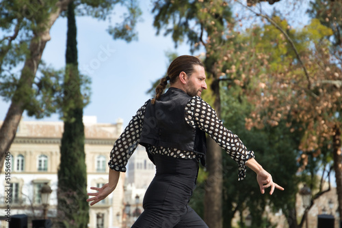 Young man with beard and ponytail, wearing black shirt with white polka dots and black pants and jacket, dancing flamenco on a stage. Concept art, dance, culture, tradition.