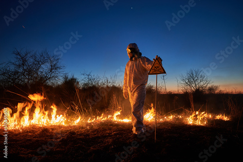 Firefighter ecologist extinguishing fire in field at night. Man in protective suit and gas mask near burning grass with smoke, holding warning sign with skull and crossbones. Natural disaster concept.