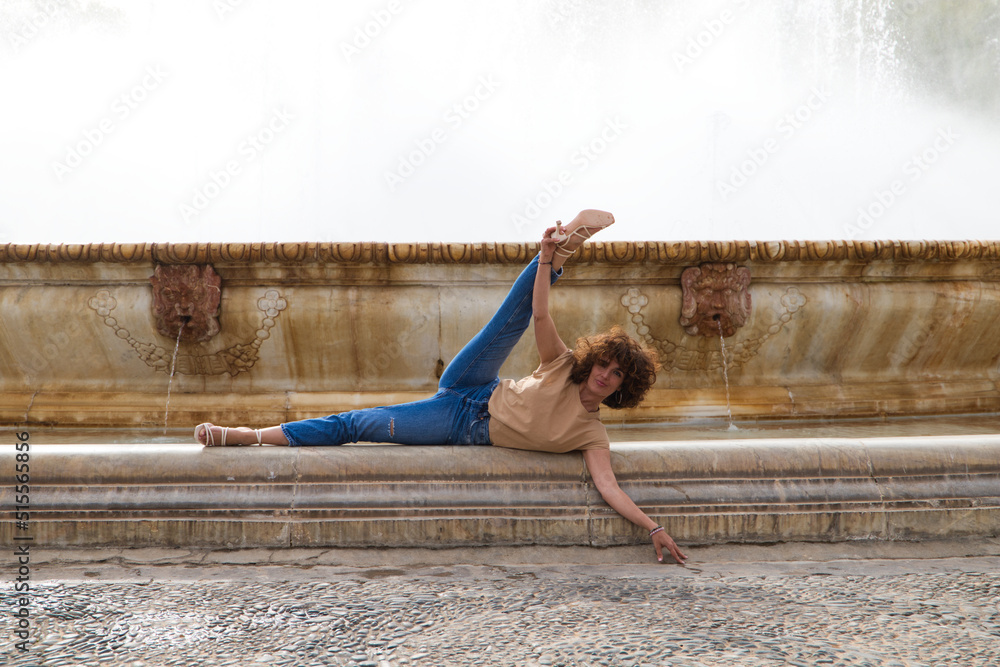 Fototapeta premium Beautiful young woman who is a dancer is standing with her legs open in a large fountain in a square. The woman is athletic and has flexibility. Well-being and health concept.
