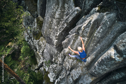 Senior alult woman climbing difficult route on a high rock with rope. Fearless climber training rock climbing on summer day. Concept of extreme sport, adventures and active lifestyle. Aerial view.