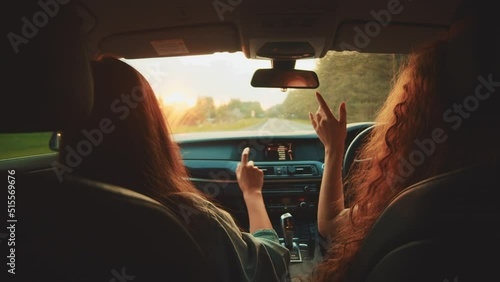 Two happy female woman friends enjoy travel in car. Sitting in front seat and have fun music song on road trip. Girls driving car and dancing. Concept of youth, friendship, holidays, sunset vacation.