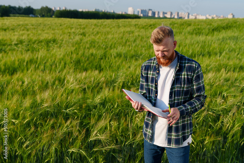 A bearded farmer with a notebook stands in a wheat field against the backdrop of a megalopolis. Preparing the field for a good ecological harvest