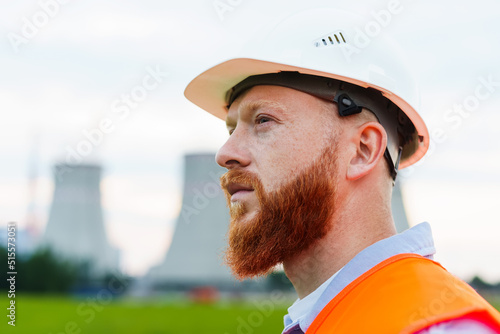 Portrait of an engineer in a white helmet and orange vest. A serious man looks at the power grid. Power plant background © makedonski2015