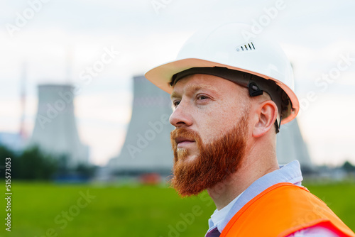 Portrait of an engineer in a white helmet and orange vest. A serious man looks at the power grid. Power plant background