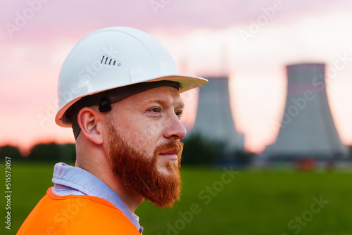 Portrait of a nuclear power plant engineer. A man with a beard in a protective helmet and an orange vest stands against the background of a nuclear power plant photo