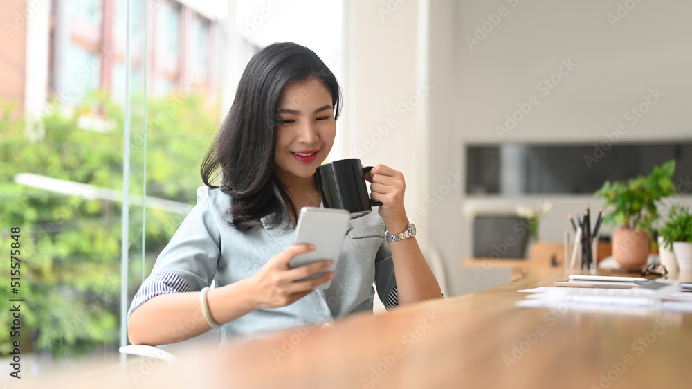 Happy young female employee sitting near office window and using smart phone Happy young female employee sitting near office window and using smart phone