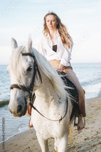 Young long hair woman in white shirt riding white horse on seascape background