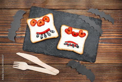 Funny monster face on halloween sandwich toast bread with butter, strawberry, cutting serving board,bats on black background close up. Kids child sweet dessert breakfast lunch food