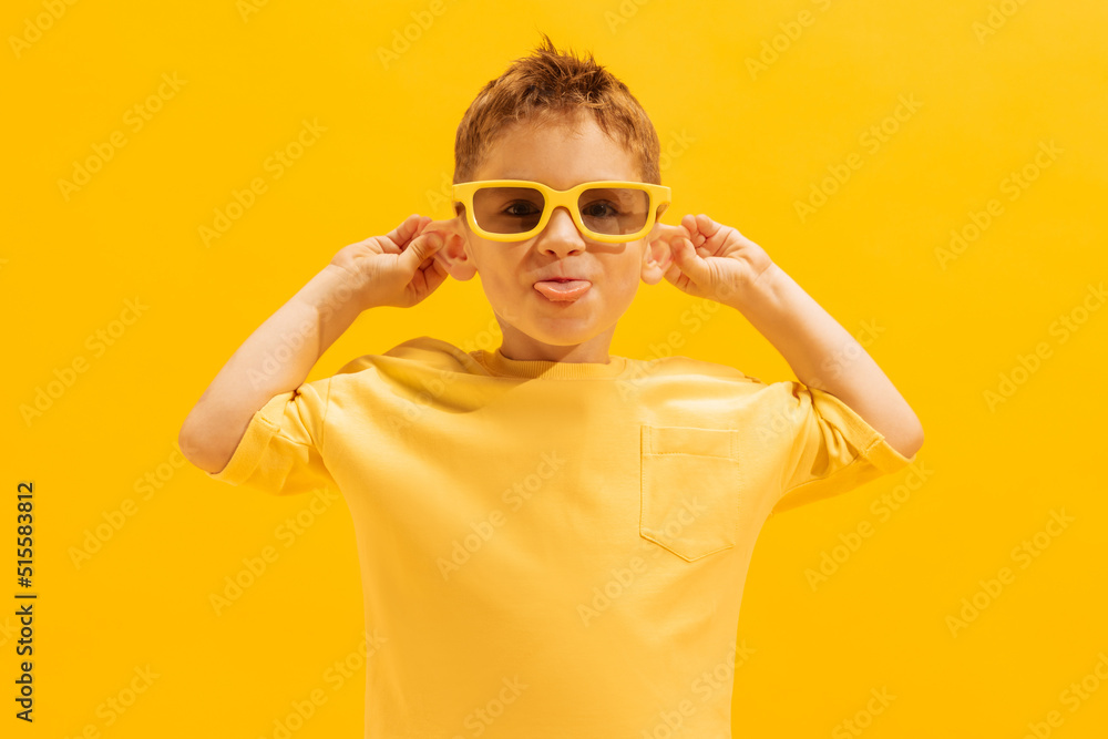 Portrait of cheerful little boy, child in bright T-shirt and sunglasses posing, making funny faces isolated over yellow background Portrait of cheerful little boy, child in bright T-shirt and sunglasses posing, making funny faces isolated over yellow background