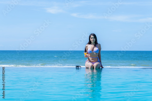 Portrait sexy asian model wearing bikini sitting on swimming pool side and drinking fruit juice in holiday summer.