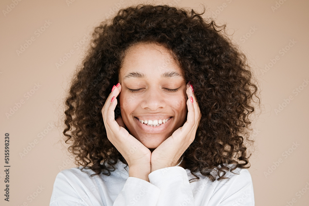 Portrait of a beautiful young woman with black curly hair. African american girl smiling end arms touch cheekbones isolated beige vivid background. People, lifestyle, beauty concept Portrait of a beautiful young woman with black curly hair. African american girl smiling end arms touch cheekbones isolated beige vivid background. People, lifestyle, beauty concept