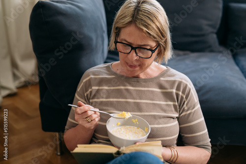 mature female reading book at home and having breakfast