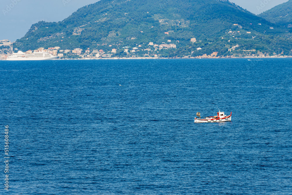 Small white and red fishing boat in the Gulf of La Spezia, Mediterranean Sea, Liguria, Italy, southern Europe. On background Porto Venere or Portovenere town and the coastline with a cruise ship.