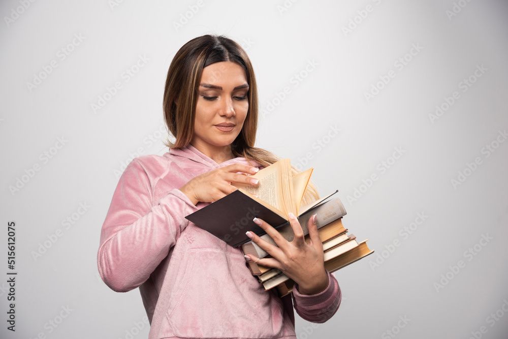 Girl in pink swaetshirt holding a stock of books, opening one on the top and reading it Girl in pink swaetshirt holding a stock of books, opening one on the top and reading it
