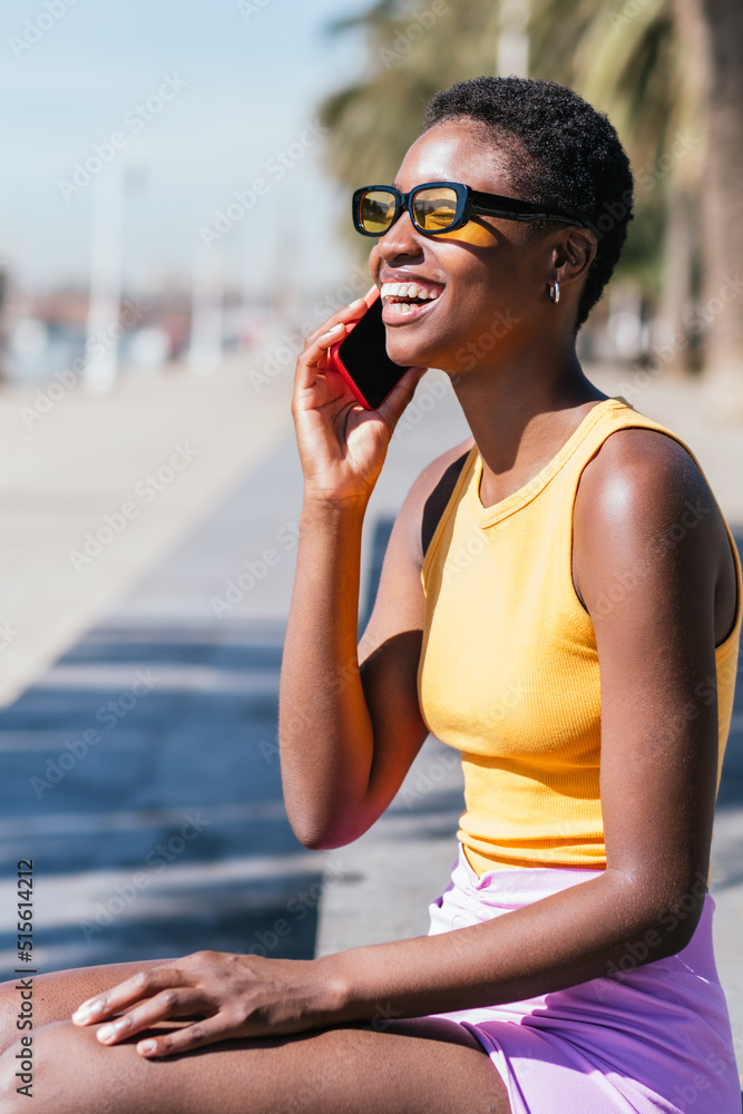 trendy african woman sitting outdoors and talking by phone. She wears casual summer attire and smiles happily