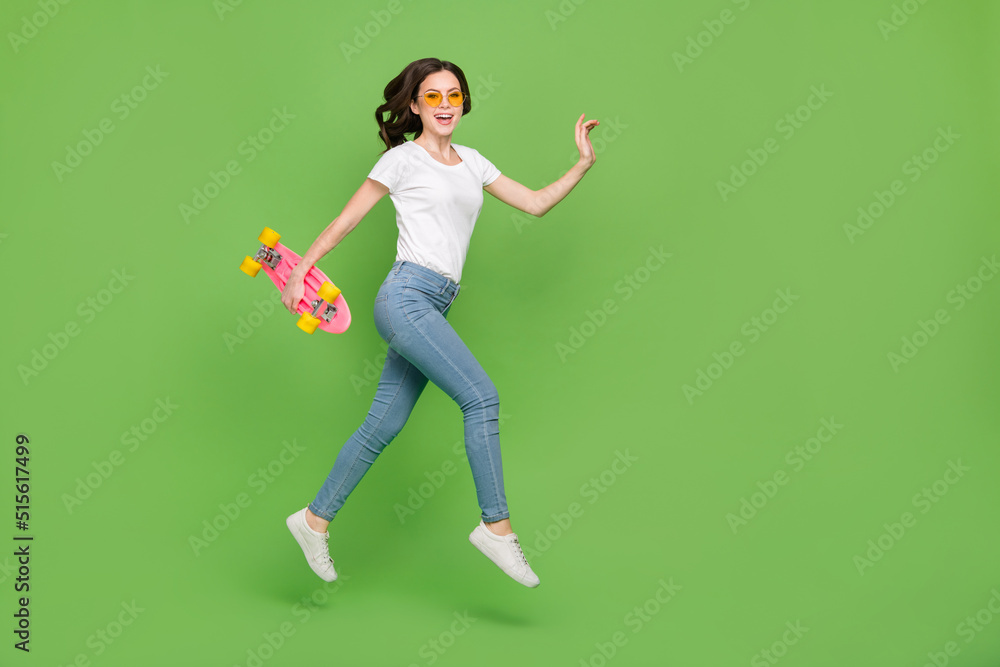 Photo of excited funny woman dressed white t-shirt eyewear holding skate board jumping empty space isolated green color background Photo of excited funny woman dressed white t-shirt eyewear holding skate board jumping empty space isolated green color background