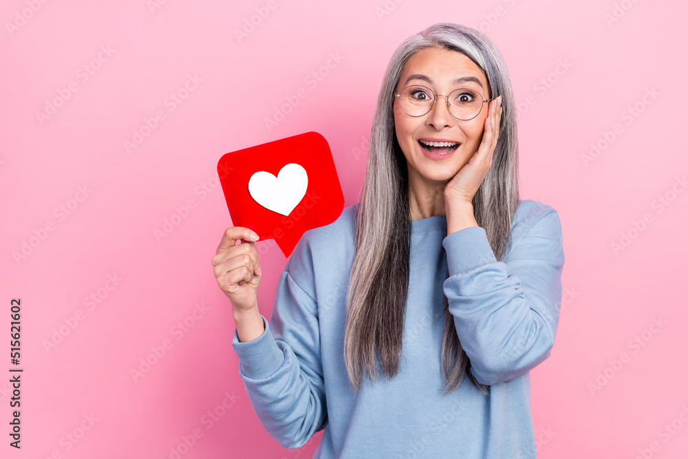 Portrait of attractive amazed impressed cheerful grey-haired woman holding like card isolated over pink pastel color background Portrait of attractive amazed impressed cheerful grey-haired woman holding like card isolated over pink pastel color background