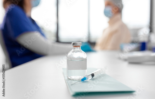 medicine, health and vaccination concept - bottle of medicine and syringe on table over doctor or nurse and patient at hospital