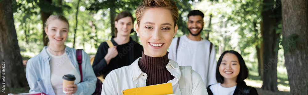 Positive student with notebook looking at camera near multicultural friends in summer park, banner. Positive student with notebook looking at camera near multicultural friends in summer park, banner.