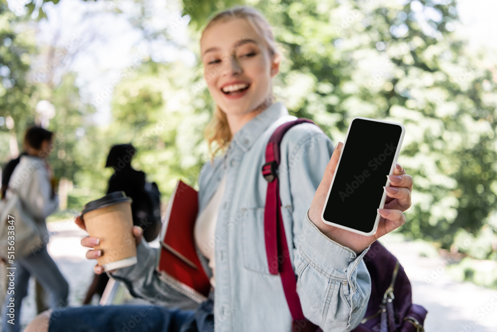 Blurred student with coffee to go showing smartphone with blank screen in park. Blurred student with coffee to go showing smartphone with blank screen in park.