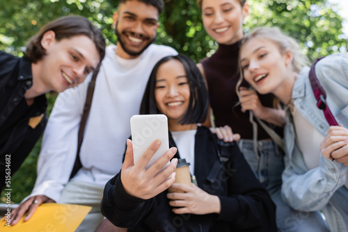 Blurred asian student holding smartphone near interracial friends in park.