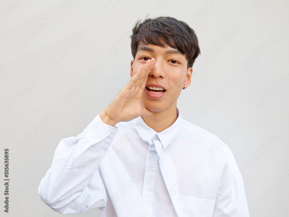 Portrait of Chinese young man with black hair in white shirt posing against white background, hand on mouth speak louder gesture, front view. Portrait of Chinese young man with black hair in white shirt posing against white background, hand on mouth speak louder gesture, front view.