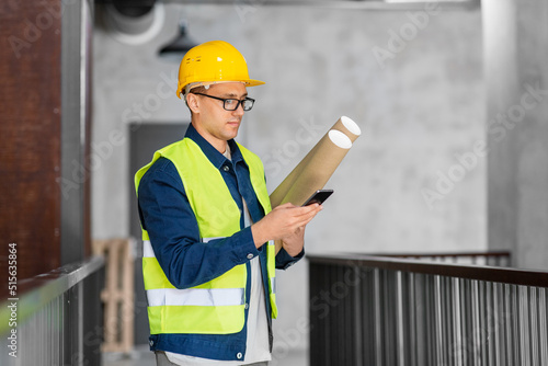 architecture, construction business and people concept - male architect in helmet with blueprint and smartphone at office