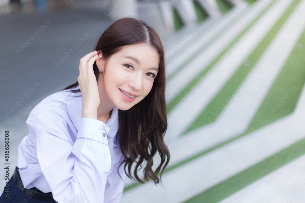 Beautiful Asian high school student girl in the school uniform with braces on her teeth stands and smiles confidently while she looks at the camera happily with the building in the background.