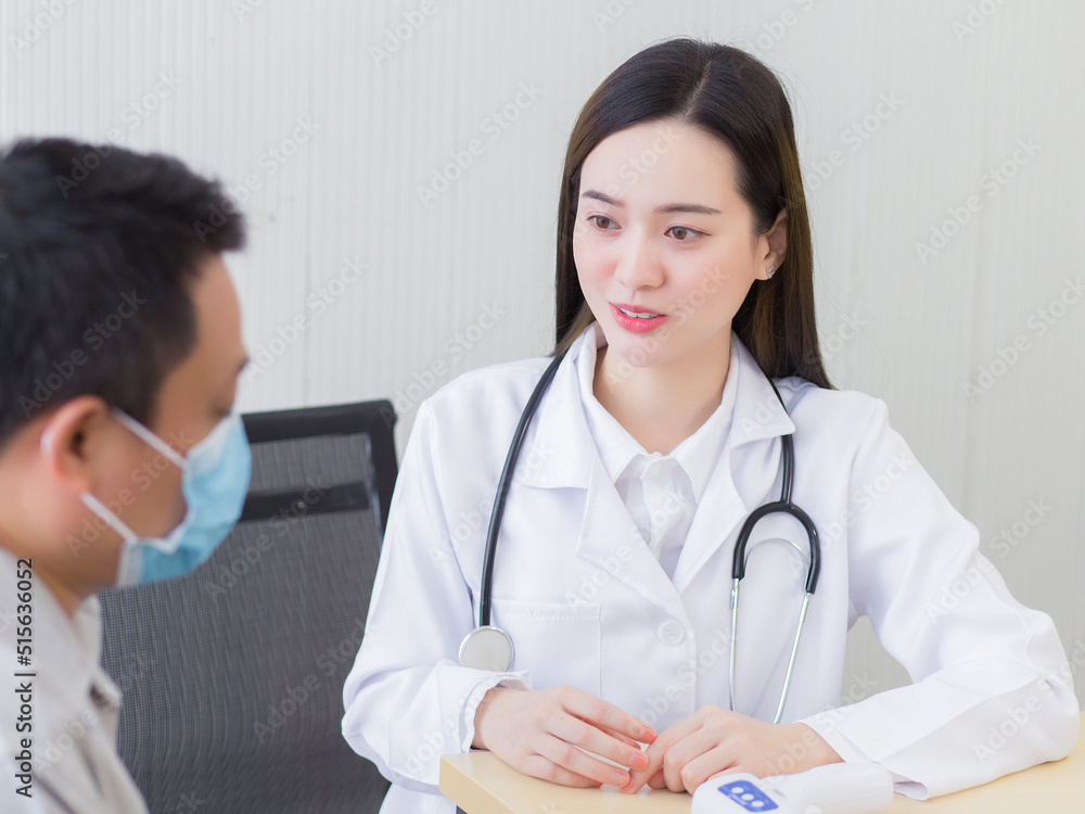 Professional beautiful young Asian female doctor talking with a man patient about his pain and symptom while they put on a face mask to Thermometer on table at the examination room in hospital.