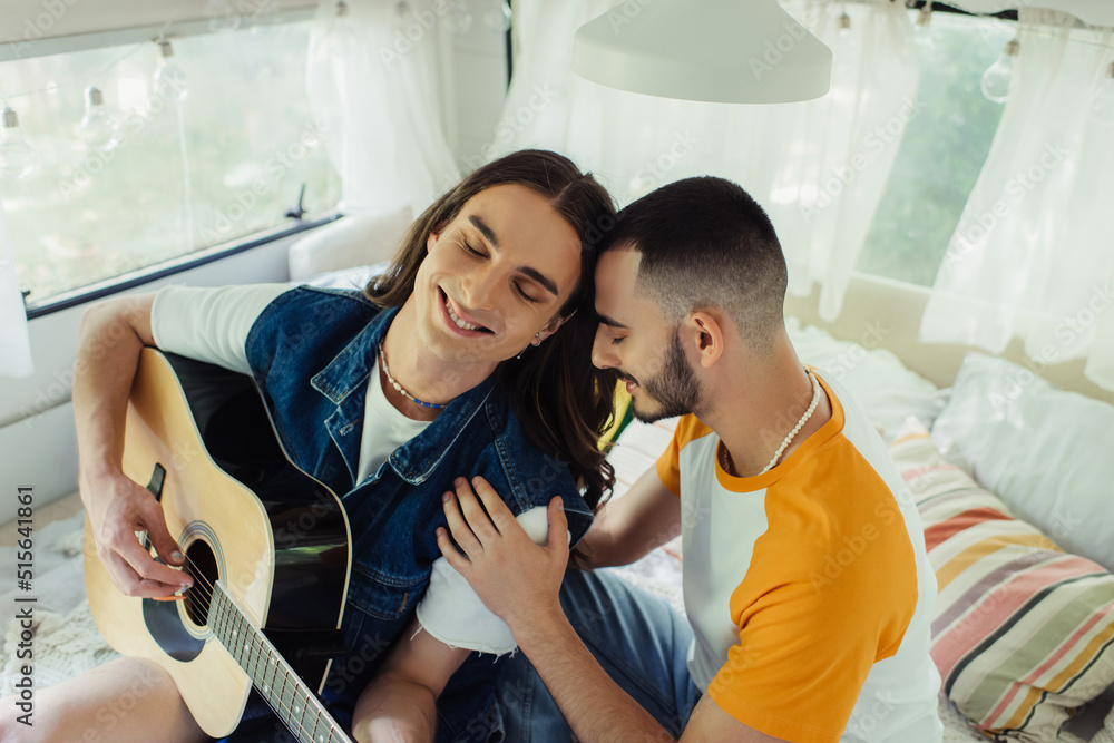 high angle view of happy gay man with long hair playing acoustic guitar near boyfriend on bed in modern van. high angle view of happy gay man with long hair playing acoustic guitar near boyfriend on bed in modern van.