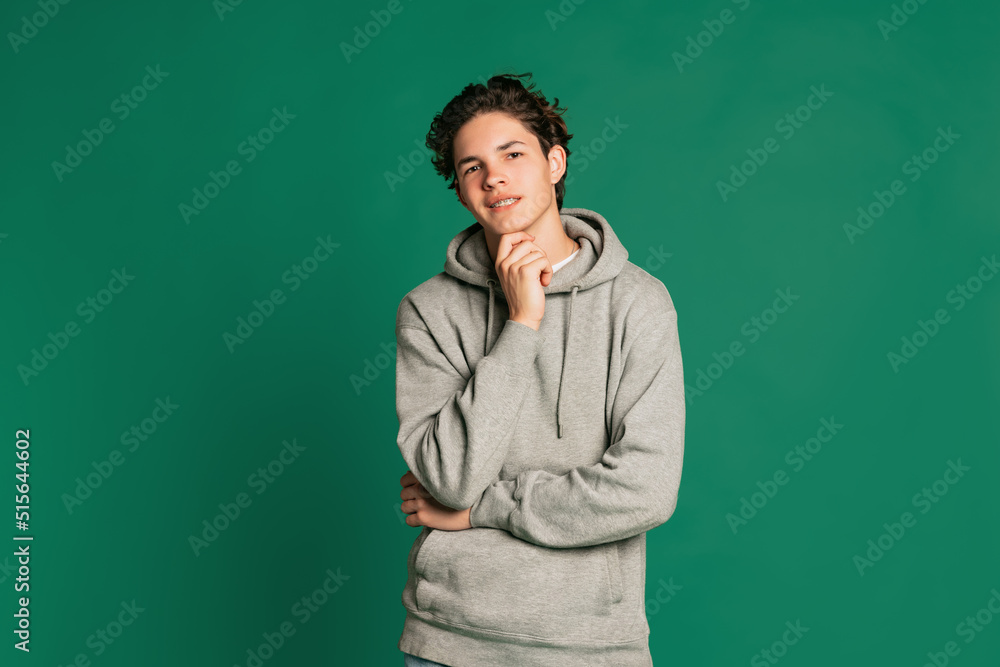 Studio shot of young boy, teen in casual style clothes looking at camera isolated over green studio background. Back to school, emotions, youth, fashion concept