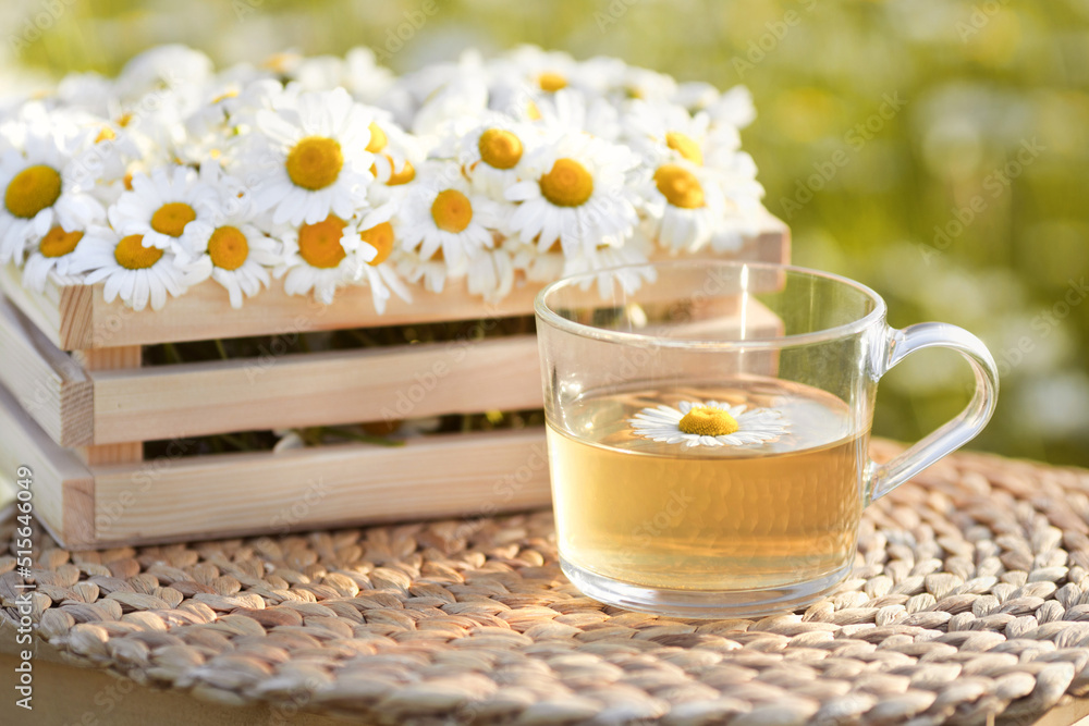 Glass cup of chamomile hot tea on background with wooden crate full of flowers outdoors