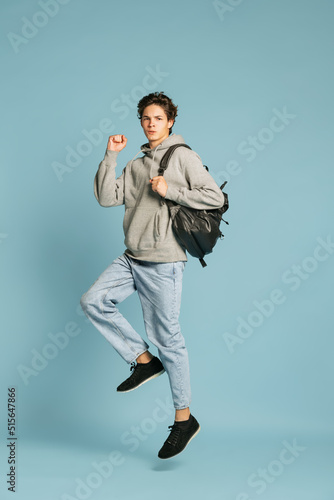 Happy student in jeans and grey hoodie jumping isolated over blue background. Concept of emotions, facial expression, youth. Back to school