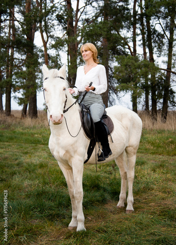 blonde beautiful woman sitting on white blue eyed  horse in green forest