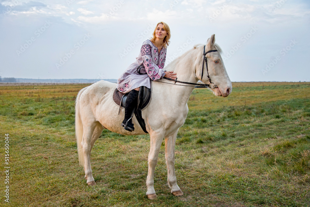 portrait of a woman in lilac ukrainian embroidery riding  white blue eyed  horse in green field