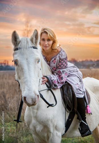 portrait of a woman in lilac ukrainian embroidery riding  white blue eyed  horse in green field © Tetatet
