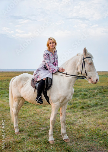 portrait of a woman in lilac ukrainian embroidery riding white blue eyed  horse in green field