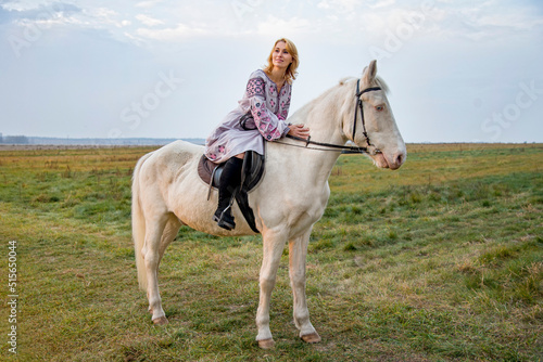 portrait of a woman in lilac ukrainian embroidery riding white blue eyed  horse in green field